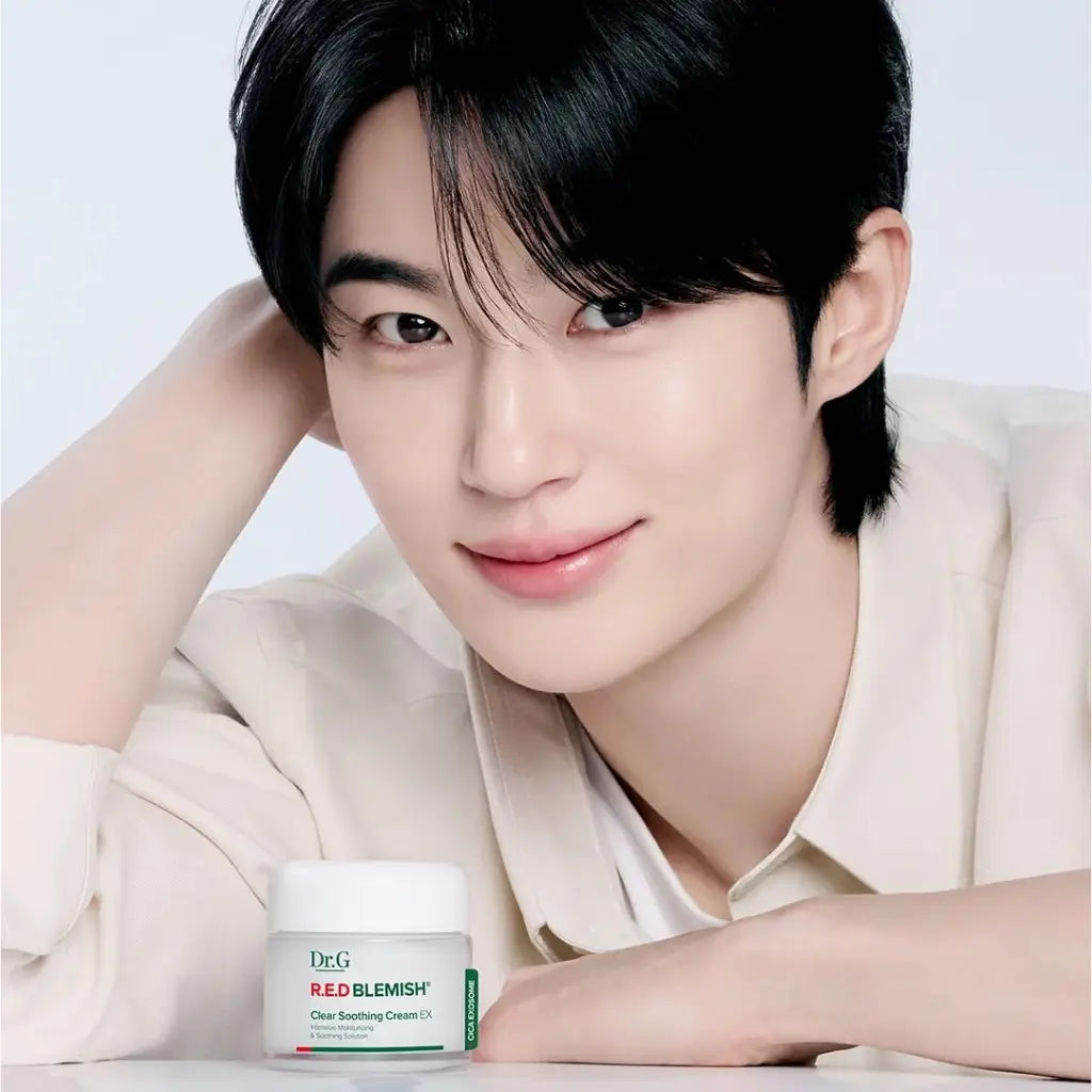 Dr.G Red Blemish Clear Soothing Cream 70ml - A smiling Korean actor man Byeon Woo-seok with dark hair rests his head on his hand, wearing a light beige shirt, with a jar of Dr.G Red Blemish Clear Soothing Cream in the foreground against a clean white background - Kbeauty Korean skincare Europe fast delivery - Skincare coréenne SKINOREA shop Paris France livraison rapide 